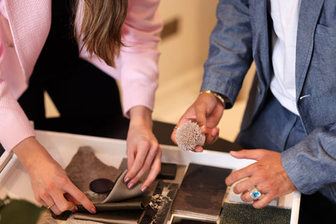 Two designers working over a fabric mood board filled with samples of fabric, wood, and marble, creating a bespoke piece of furniture. The designers are selecting materials for a unique and personalized sofa, armchair or chaise lounge, with a focus on quality and style.