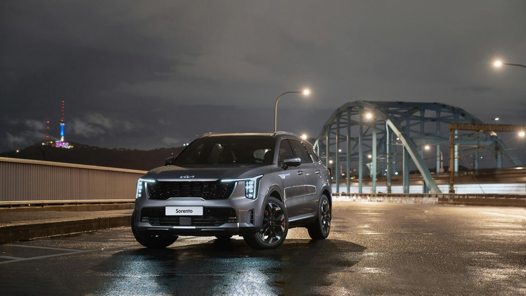 Silver Kia Sorento SUV parked on wet road at night with illuminated bridge and city lights in the background