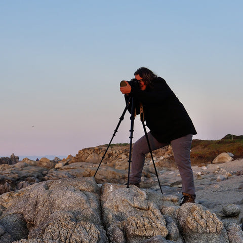 A woman with a camera on a tripod atop on rocks at the shore.