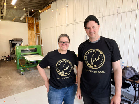 Two people in a puzzle workshop looking at the camera in a friendly way and wearing black T-shirts featuring the Oakland Puzzle Company logo.