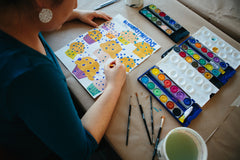 A woman with watercolor paints working on a piece showing muffins with the words Blueberry Muffins