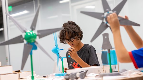 The image displays a moment in an educational setting, which appears to be a science classroom or workshop, where children participate in a hands-on project involving wind turbines. In the foreground, a child with glasses intently examines a small piece, which seems to be a component of the turbine model. In the background, other children can be seen enthusiastically holding up assembled wind turbine models. The wind turbines have a green central hub with blue stands and gray blades. The setting suggests a learning environment encouraging interactive and practical understanding of renewable energy concepts. The boxes and materials on the table indicate that the activity might involve assembling or customizing these models as a part of their learning experience.