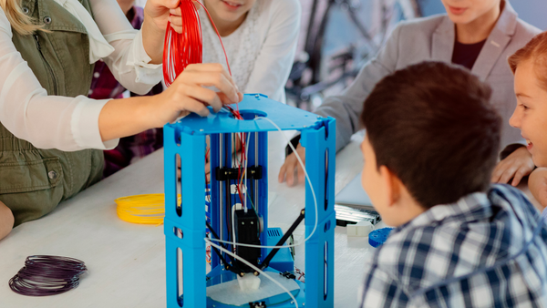 This picture depicts a group of children participating in an educational activity, perhaps a science or technology class. The focus is on a 3D printer, which has a blue frame. One of the children is actively interacting with the printer, possibly loading or adjusting filament. Filament spools in purple and yellow are resting on the table, indicating that the children can choose the colors for their projects. The children appear curious and attentive, suggesting they have a hands-on learning experience involving technology and engineering. The bright and collaborative environment indicates that the students are taught through a modern approach that fosters creativity and practical skills.