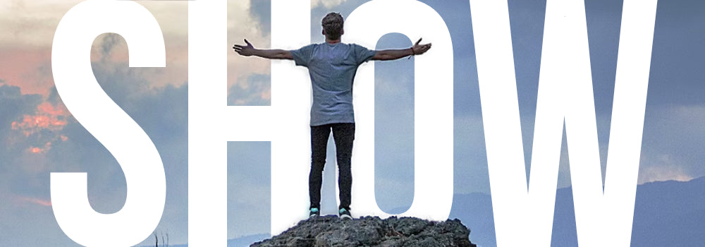Un hombre que extiende sus brazos con valentía en la cima de una montaña con la palabra "Show" detrás, que representa camisetas cristianas que muestran su fe.