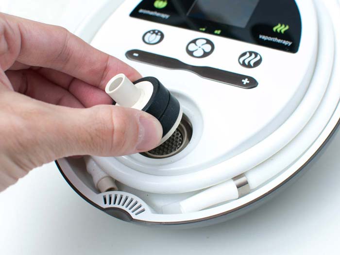 The image shows a hand inserting a white and black silicone or rubber fitting into the central heating element of a Herbalizer vaporizer. The top of the Herbalizer with its touch controls for aromatherapy and vaportherapy, along with temperature adjustment buttons, is clearly visible. The white silicone whip attachment is coiled around the base of the device. The hand is carefully aligning the fitting with the heating chamber.