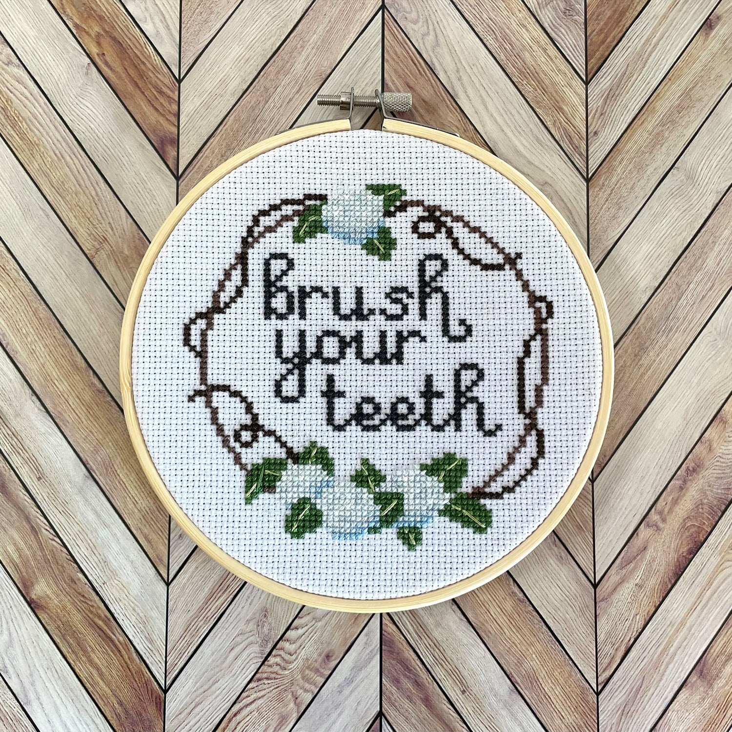 A finished cross-stitch in an embroidery hoop. The design of the cross-stitch reads "brush you teeth" and has a floral border decoration.