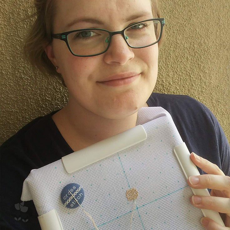 A digital photograph of grown-up Kate, standing in front of a yellow-ish, textured wall. She now wears glasses and has a sombre smile whilst looking at the camera. In the one hand you can see, she’s holding a q-snap with Aida cloth stretched in it, where she’s begun working on a stitch people portrait.