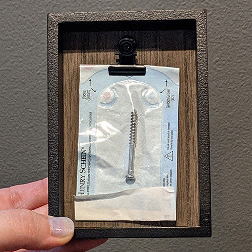 A picture of a surgical screw in a sterile packet that is clipped into a little shadowbox frame.