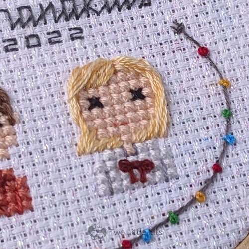 A close-up photo of a cross-stitched little         blonde-haired girl. She is wearing a top that has a red bow tied on the front and you can also see a string of hand embroidered 'Christmas lights' around her.