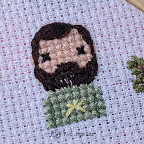 A close-up photo of a cross-stitched man. He has lots of dark brown facial hair that, at the chin -only, is speckled with salt &amp; pepper colouring. He is wearing a light green top with a yellow star on the front.