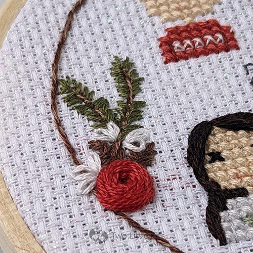 A close-up photo of hand-embroidered red flower that has sprigs of pine, some white flowers and pinecones. Underneath you can see the curve of a simple brown circular border.