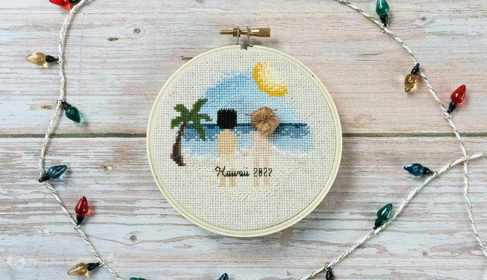 alt="A flat-lay photograph of a cross-stitch couple portrait in a 4in embroidery hoop. The stitch depicts a man and woman standing on a beach (with palmtree and sun in the design) however, they are both naked. You can just make out underwear tan lines and butt-cracks. The woman's hair is stitched up to look like a bun. Around the hoop is a string of fake christmas lights."