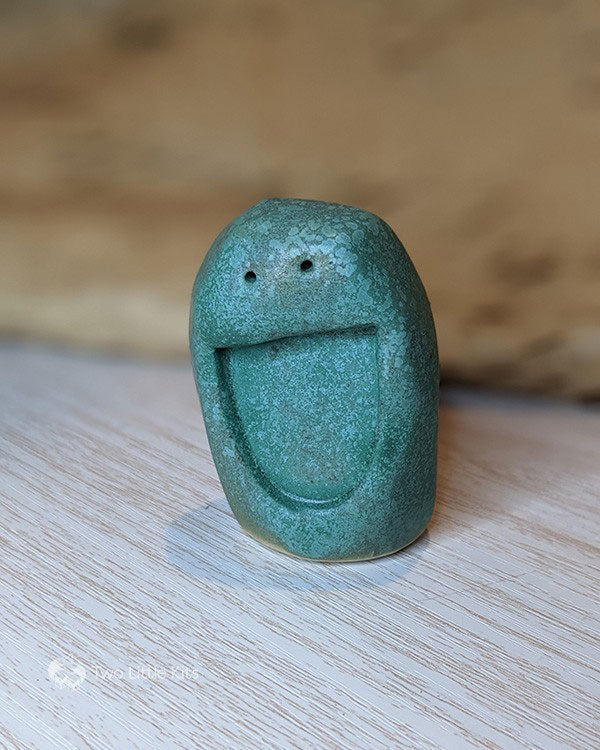 A green-glazed clay, tall 'blob' which is about the length of your palm, that's been stamped (before fired) with a semi-circle mouth and two tiny little circle eyes, so it looks like it has an enormous smiley face. The green glaze is mottled in appearance.