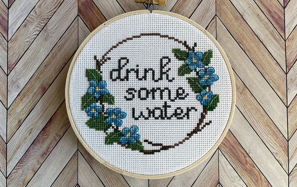A finished cross-stitch in an embroidery hoop. The design of the cross-stitch reads "Drink some water" and has a floral border decoration.