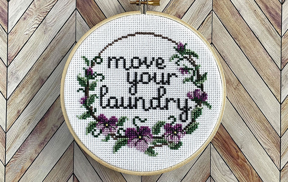 A finished cross-stitch in an embroidery hoop. The design of the cross-stitch reads "Move your laundry" and has a floral border decoration.