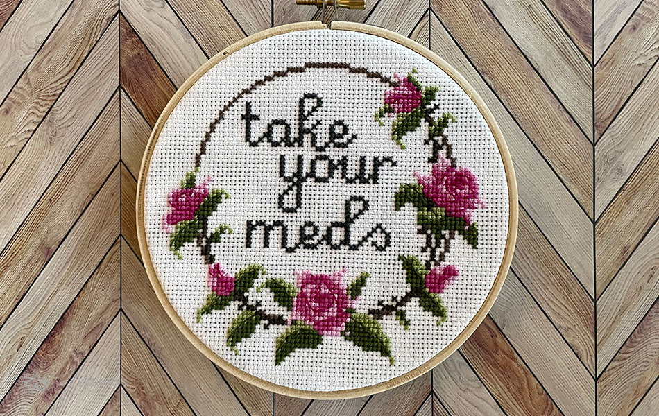 A finished cross-stitch in an embroidery hoop. The design of the cross-stitch reads "Take your meds" and has a floral border decoration.