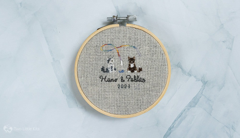 A 4" embroidery hoop depicting two cross-stitched cats with some cat toys. It says "Hans & Pablito 2023" underneath them.