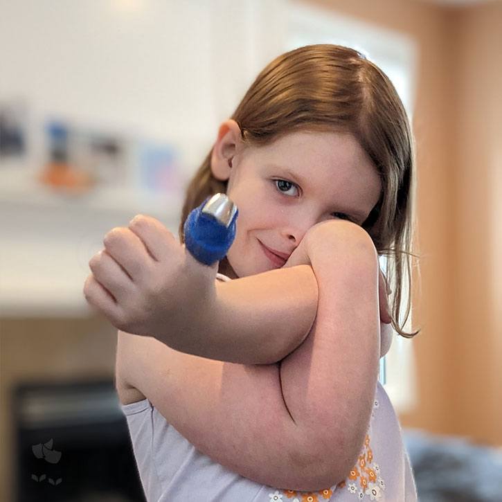 A young girl partially covering her face with her arms. She is sticking out her left hand giving a thumbs up, however her thumb is wrapped up in a splint and bandage.