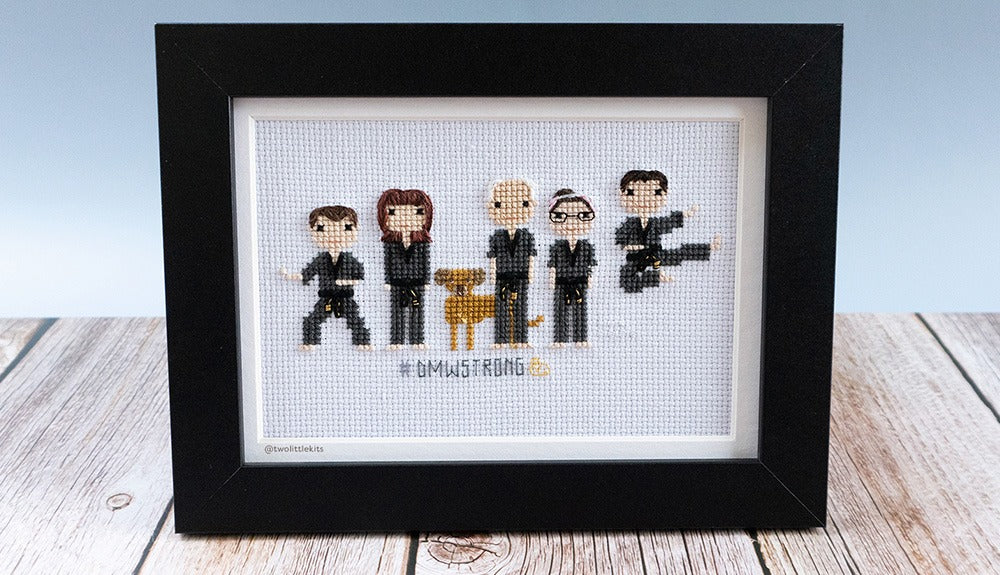 A photo of a framed, completed cross-stitch piece that depicts 5 people and 1 dog. The people are all in black martial arts uniform, two of them are in 'motion' in a way. The dog that is shown is a brown colour and is standing behind the man in the middle.