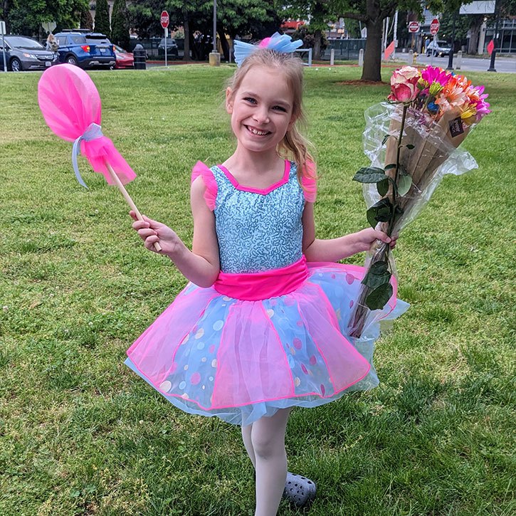 A young girl in a blue and pink dress with a bouquet of flowers and a pretend pink lollipop