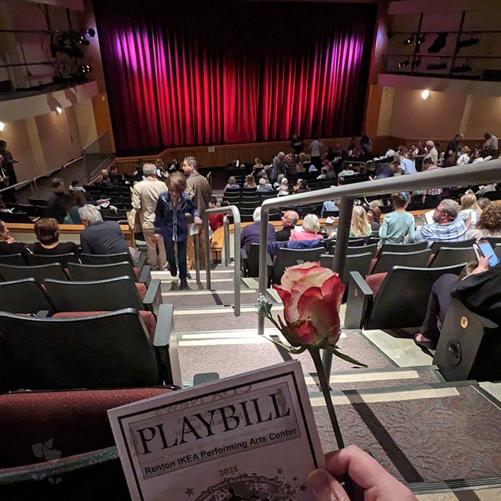 Photo of Kate holding a playbill and a single rose at the back of a full auditorium before the show starts.