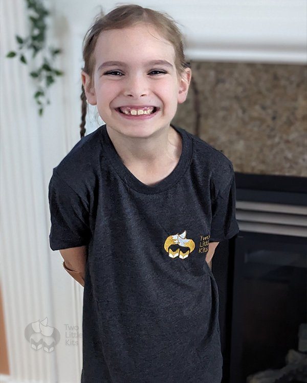 A young girl smiling while wearing a 'Two Little Kits' t-shirt.