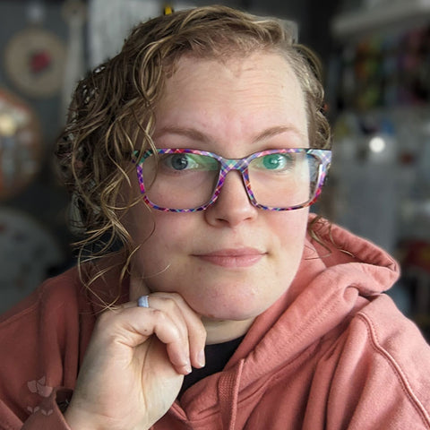 Kate gently smiling at the camera. She's wearing glasses that have a magnetic topper that change the frames to be a "berry" plaid colouring. Her hair is curly and she's wearing a peach-coloured, cosy hoodie.