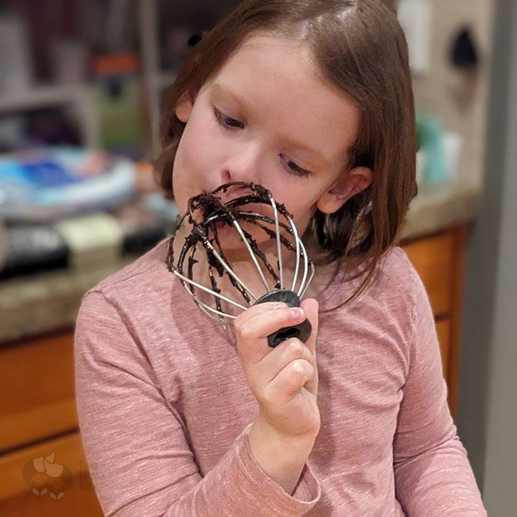 A little girl enjoying licking a mixing beater that has leftover brownie mixture on it.