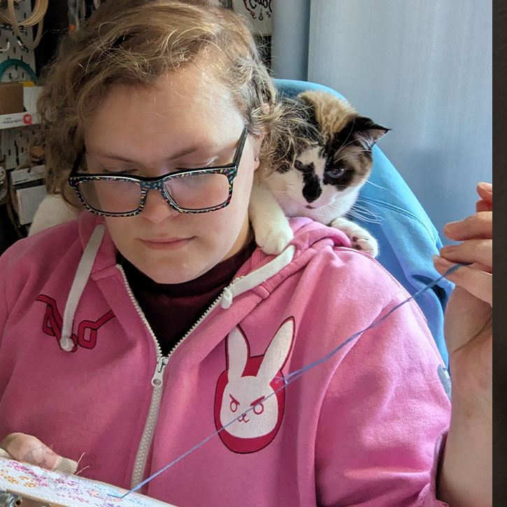 A cat sitting on a woman's shoulders while she cross-stitches