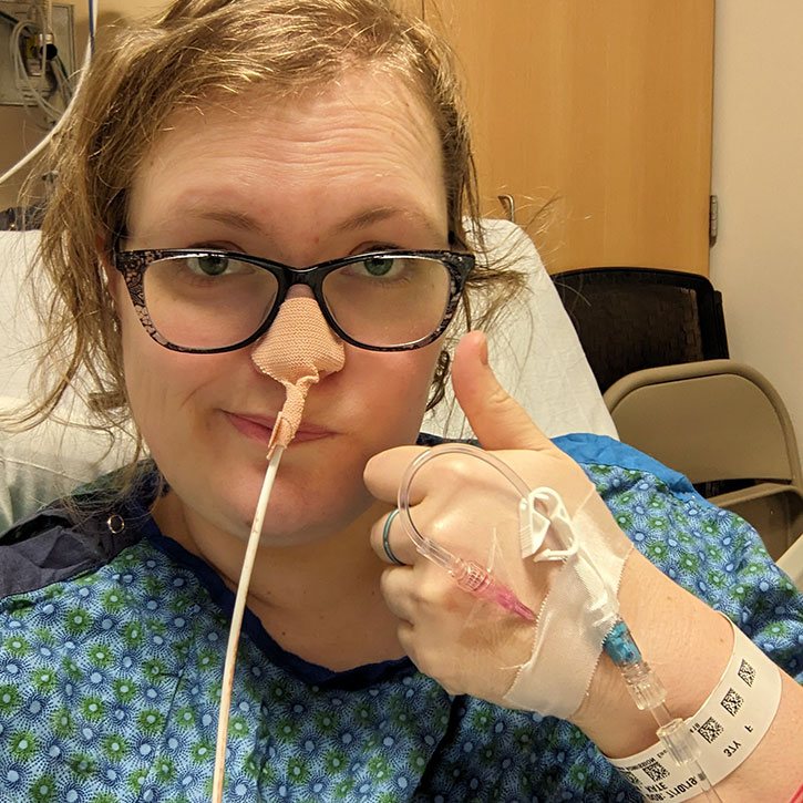 A disgruntled Kate giving a (sarcastic) thumbs up. She has a feeding tube coming out of her nostril and she's wearing a hospital gown.