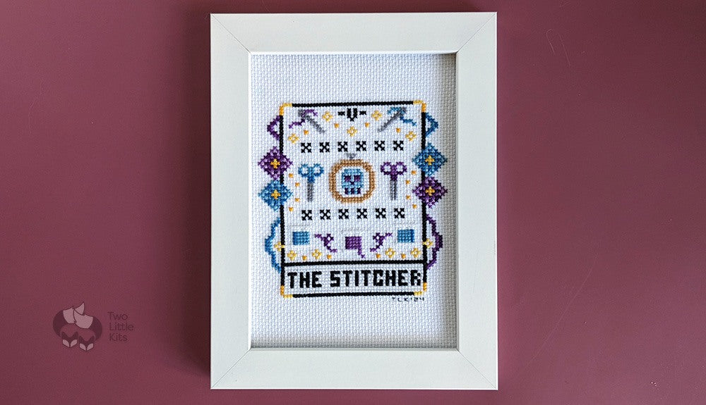 A finished, framed cross-stitch piece of a pretend tarot card called "The Stitcher". The imagery shows stitchy paraphernalia on and around it.