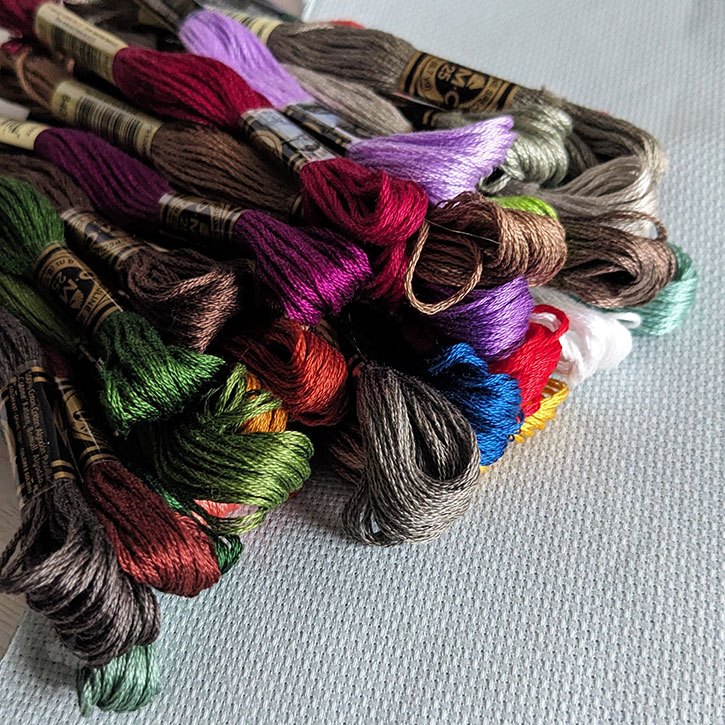 A bundle of DMC floss skeins laying in a pile on top of a piece of Aida fabric