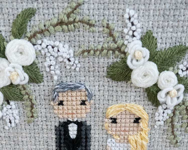 A cross-stitch piece depicting the tops of a bride and groom, with hand embroidered, white florals above and beside them. The photo is up-close to the piece itself.