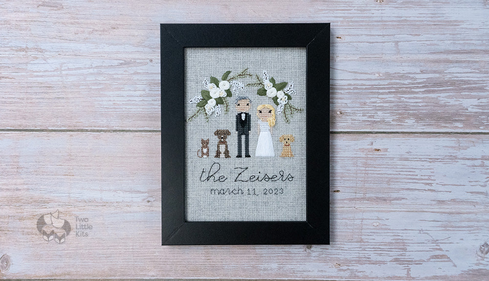 A wedding portrait done as a cross-stitch & hand embroidered piece of artwork. It's framed in a simple, black 5x7" frame and the piece depicts a bride & groom alongside their two dogs and one cat. There is floral decorations above the characters.