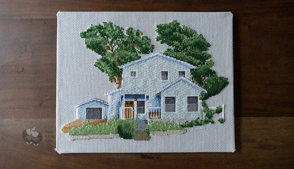 A large, 8"x10" cross-stitched house piece. The home is a blue colour with a front porch and two stories, with a detached garage. There are two large trees towering above behind the home and some greenery out the front.