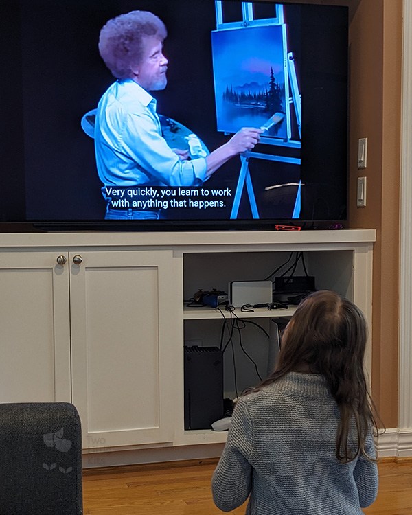 A young girl watches TV with a video of painter Bob Ross on it