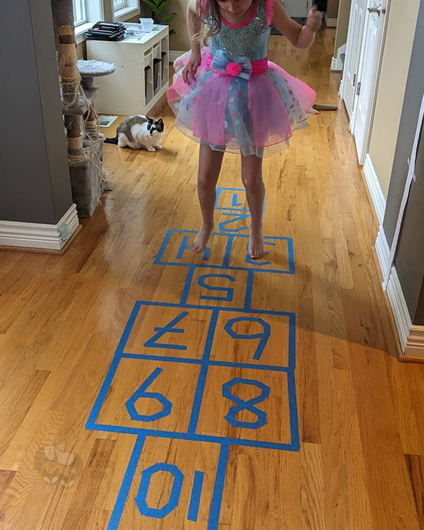 A hopscotch design made with blue painters tape on a hardwood floor. A girl in a pink & blue dress is actively doing it