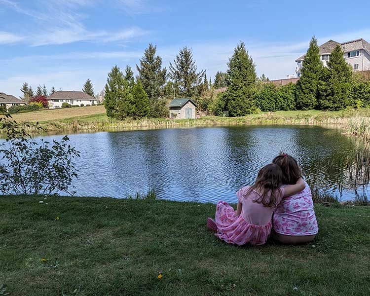 Two young girls sit together, cuddling while looking out over a pond.