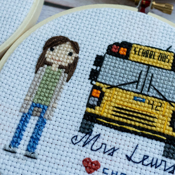 A close-up, angled photograph of a finished cross-stitch piece that depicts a school bus driver character alongside their school bus.
