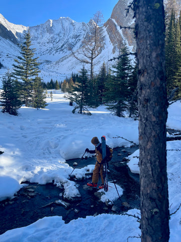 Kid carrying skis across a river while backcountry skiing.