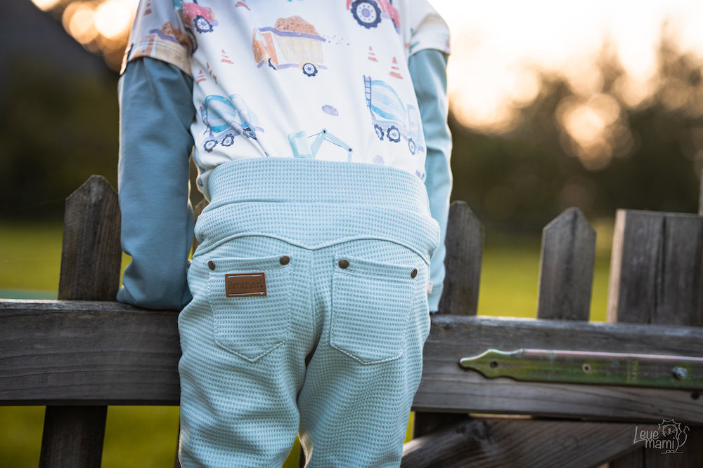 Kleiner Junge steht auf einer Wiese im Sonnenuntergang. Er trägt selbstgenähte Kleidung aus Biostoffen von Faybee. Das Bild zeigt den Jungen von hinten. Die Hose ist aus einem tollen Waffelstrick genäht.