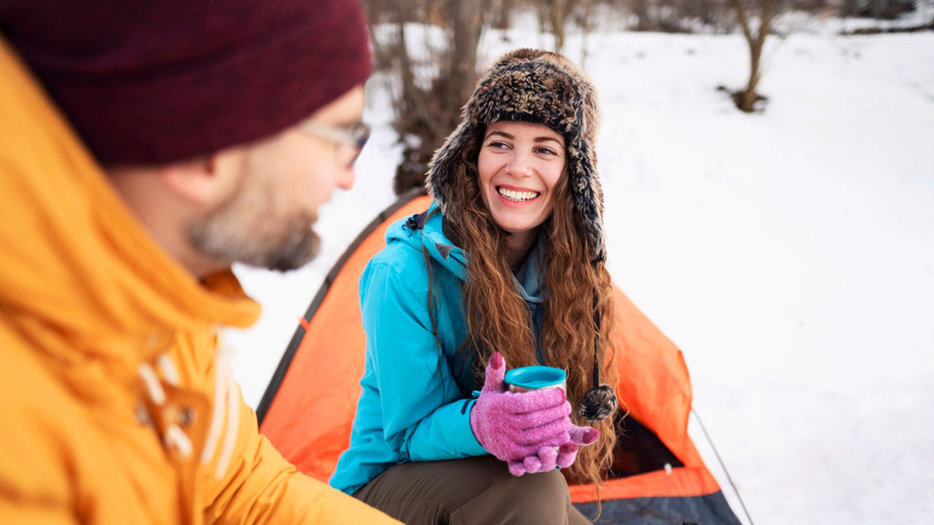 Essential winter camping gear. Happy couple enjoying coffee at campsite in snow. I Go Overland Blog.