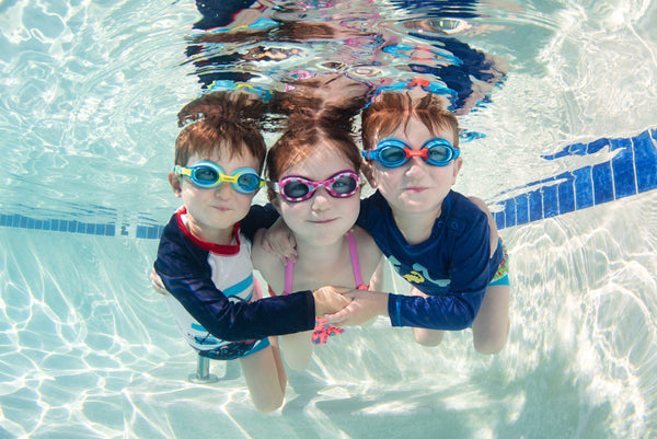 Three little kids swimming underwater wearing colorful goggles and smiling.