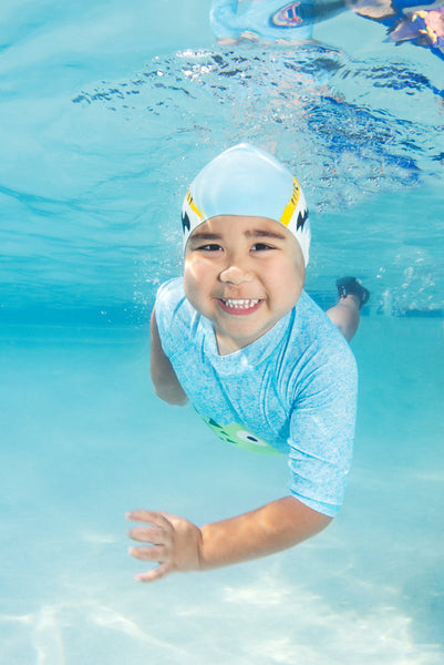 Little boy swimming underwater wearing a blue swim cap and smiling.