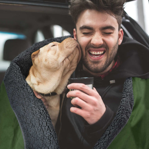 A man in a car draped in a green waterproof blanket with a dog kissing his face next to him
