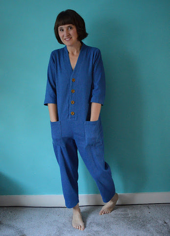 Zoe Edwards standing against a bright blue wall, wearing a blue overall, bare feet, hands in pockets