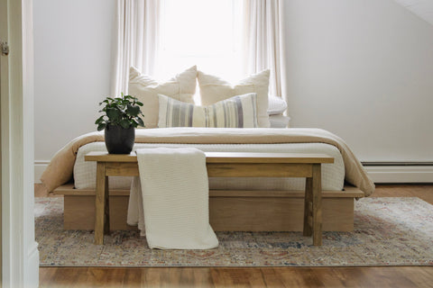 Wooden bench sitting in front of a bed. The bench is styled with a green plant and a white throw blanket.