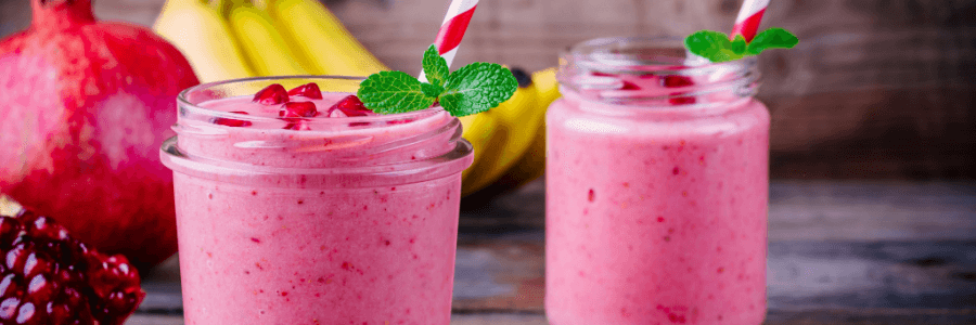 two pink smoothies in glasses with pomegranate and banana in background