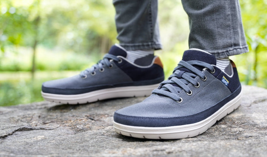 Diagonal view of a person wearing a pair of Lems Chillum Varsity Blue shoes standing on a flat stone with greenery in the background