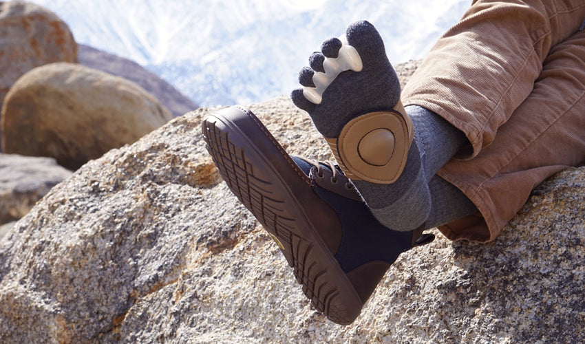 A hiker, sitting on a rock, wearing Correct Toes, Strutz foot pads, Injinji toe socks, and Lems Boulder Boots